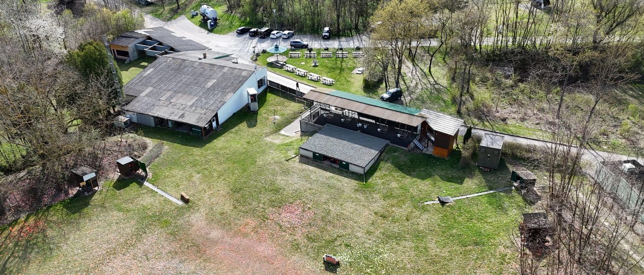 Eine Luftaufnahme eines Bauernhofs mit mehreren Gebäuden, einem Parkplatz und einem Grasfeld. Bäume und Sträucher umgeben das Gebiet.