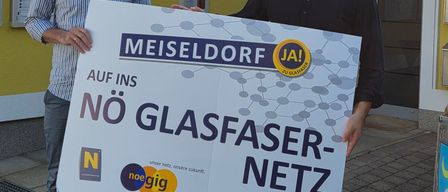 Zwei Männer stehen vor einem Gebäude und halten ein großes Schild mit den Worten Meiseldorf und Auf ins No Glasfaser-Netz. Sie lächeln.