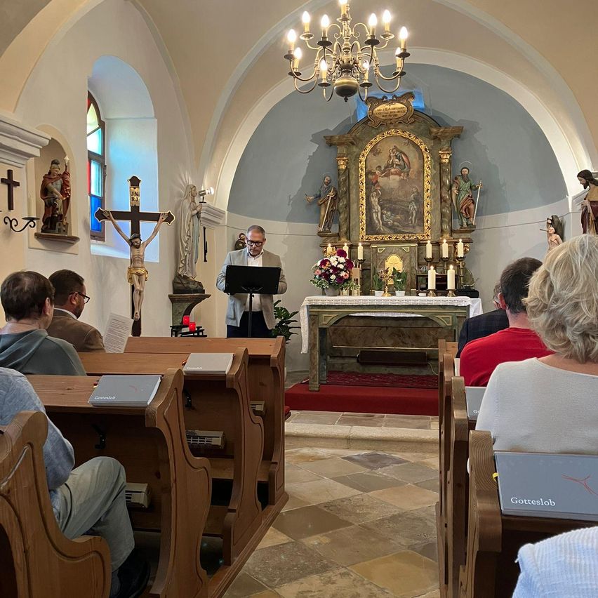Ein Mann steht an einem Podest in einer Kirche und spricht zu einer Gemeinde. Der Altar hat ein Gemälde, Kerzen und Blumen. Bänke sind mit Menschen gefüllt, einige mit Büchern. Eine Kronleuchter hängt über ihnen.