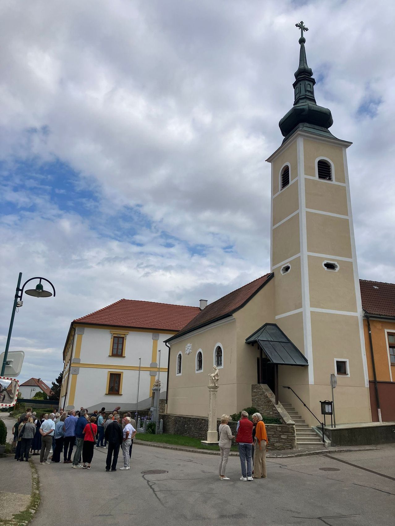 Eine Gruppe von Menschen versammelt sich vor einer Kirche mit Turm unter einem bewölkten Himmel.