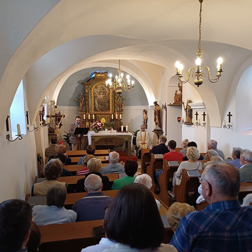 Eine Gruppe von Menschen nimmt an einem Gottesdienst teil. Ein Priester steht am Altar. Die Gemeinde sitzt auf Kirchenbänken, und die Kirche ist mit Kerzen und religiösen Statuen geschmückt.