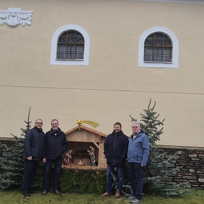 Vier Männer stehen vor einer Krippe vor einem beigen Gebäude mit bogenförmigen Fenstern.