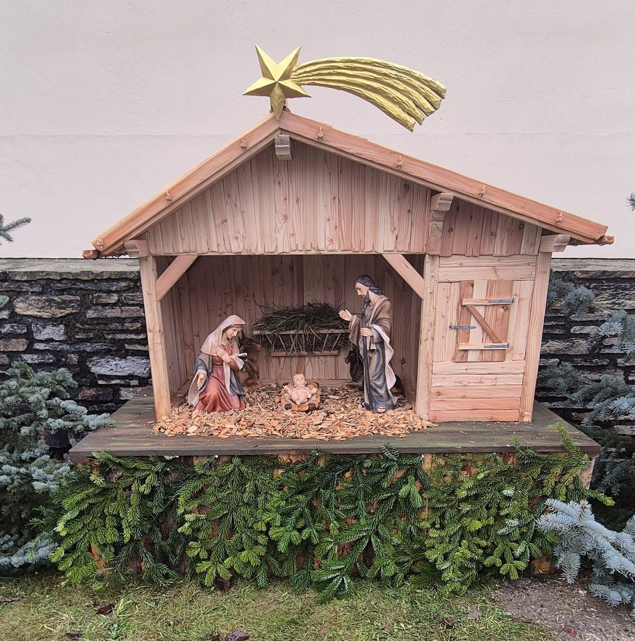 Eine Krippe ist vor einem Gebäude aufgestellt und zeigt Maria, Josef und das Jesuskind in einer hölzernen Krippe. Ein goldener Stern schmückt das Dach.