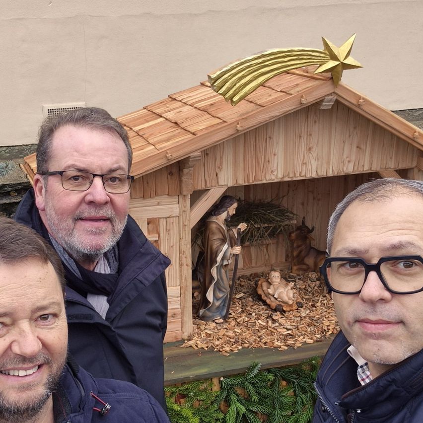 Drei Männer lächeln vor einer hölzernen Krippe mit einem goldenen Stern. Der Mann links hat einen Bart, und die anderen tragen Brillen.