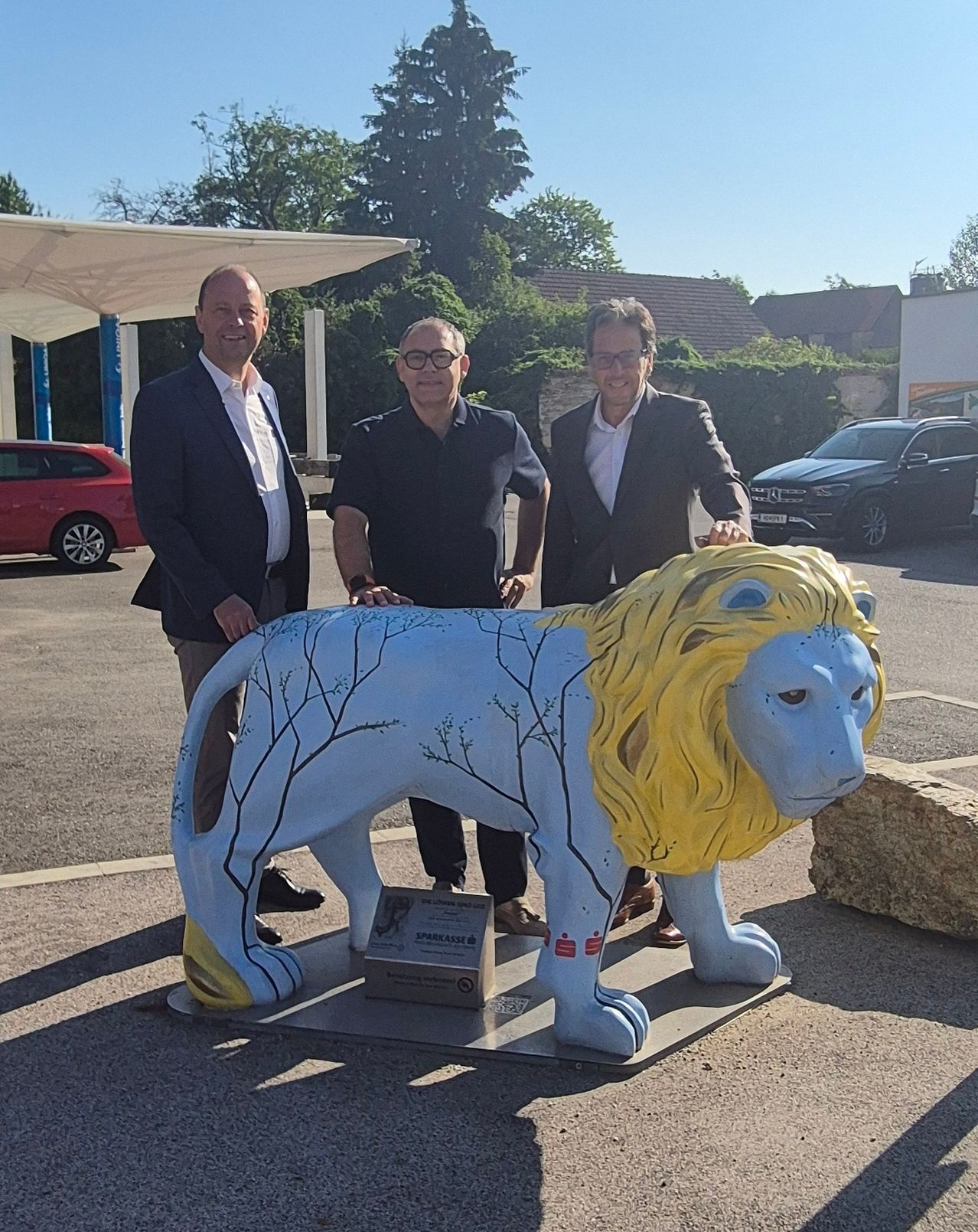 Drei Männer stehen neben einer großen bemalten Löwenstatue auf einem Parkplatz mit einem roten Auto und SUV im Hintergrund.