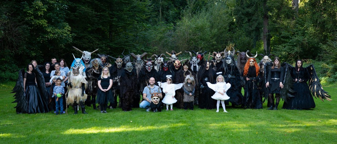 Eine Gruppe von Menschen in Kostümen, einschließlich Engel und Teufel, posiert für ein Foto in einem Grasbereich mit Bäumen im Hintergrund.