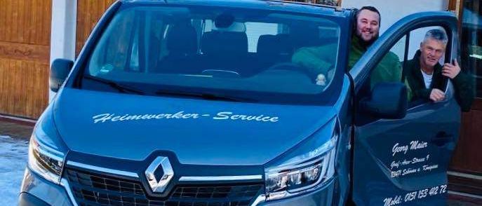 Zwei Männer stehen neben einem blauen Renault-Van, die Fahrertür ist offen. Sie lächeln und einer hält die Tür. Der Van hat 'Heimwerker-Service' auf der Seite geschrieben.