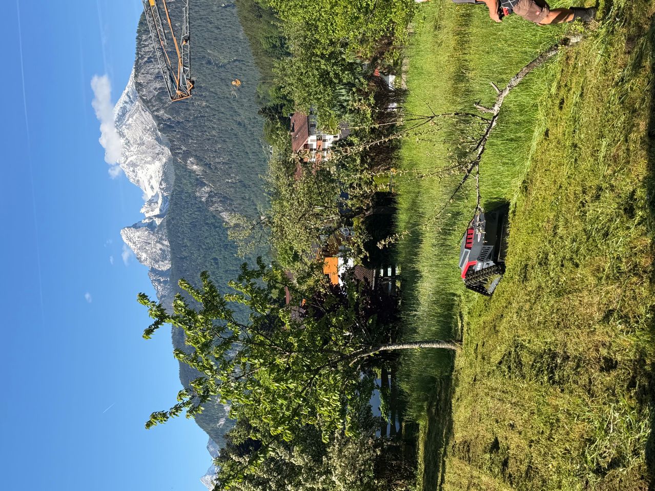 An aerial view shows a mountainous landscape with a crane and a person in a grassy field. The scene includes a house and a snowy mountain peak.