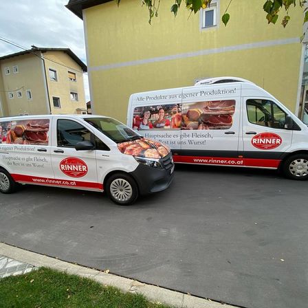 Zwei mit Werbung für 'Rinner' beklebte Lieferwagen stehen vor einem gelben Gebäude. Die Lieferwagen haben Bilder von Fleischprodukten und den Slogan 'Hauptsache es gibt Fleisch, der Rest ist uns Wurst!' darauf.