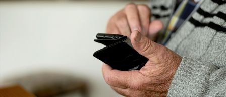 Eine ältere Person hält einen schwarzen Geldbeutel in den Händen, der Hintergrund ist verschwommen, möglicherweise drinnen.