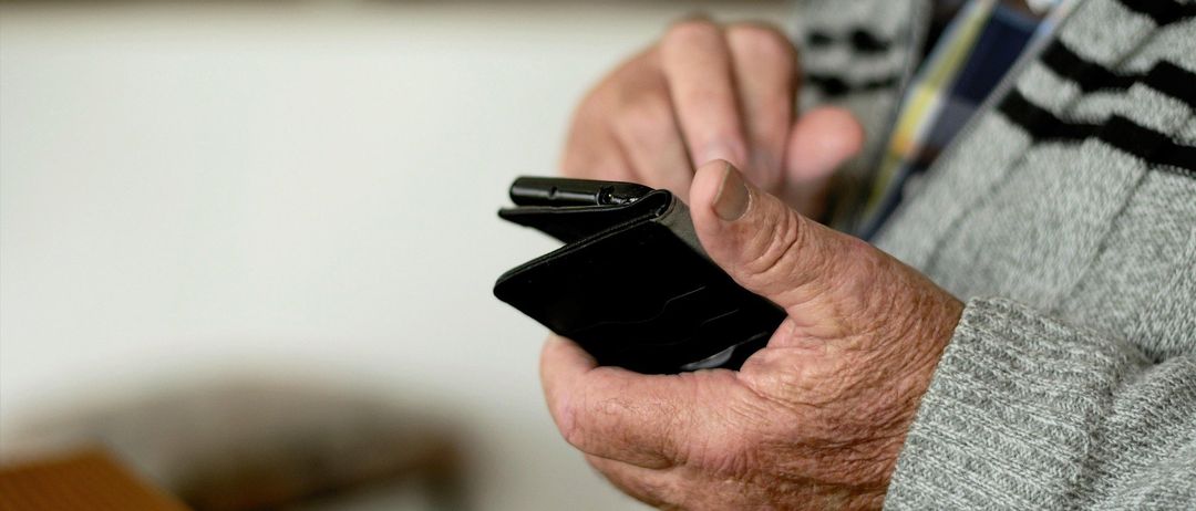 Eine ältere Person hält einen schwarzen Geldbeutel in den Händen, der Hintergrund ist verschwommen, möglicherweise drinnen.