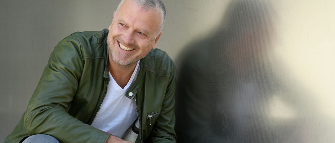 Ein Mann in einer grünen Lederjacke und weißem T-Shirt, sitzt auf einer Metallwand, lächelt und zeigt mit der Hand.