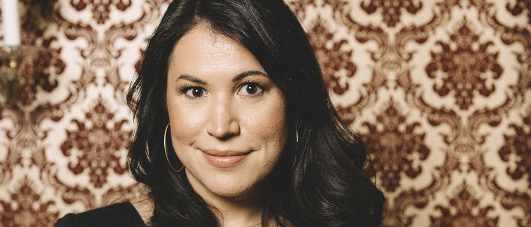 Eine Frau mit schwarzem Haar und einem warmen Lächeln steht vor einer Tapete mit einem Blumenmuster. Sie trägt ein schwarzes Oberteil und Creolen.