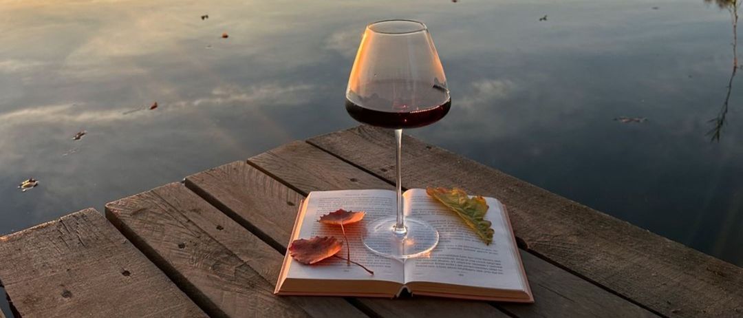 Ein mit rotem Wein gefülltes Weinglas steht auf einem offenen Buch auf einer Holzterrasse mit Blick auf einen ruhigen See.
