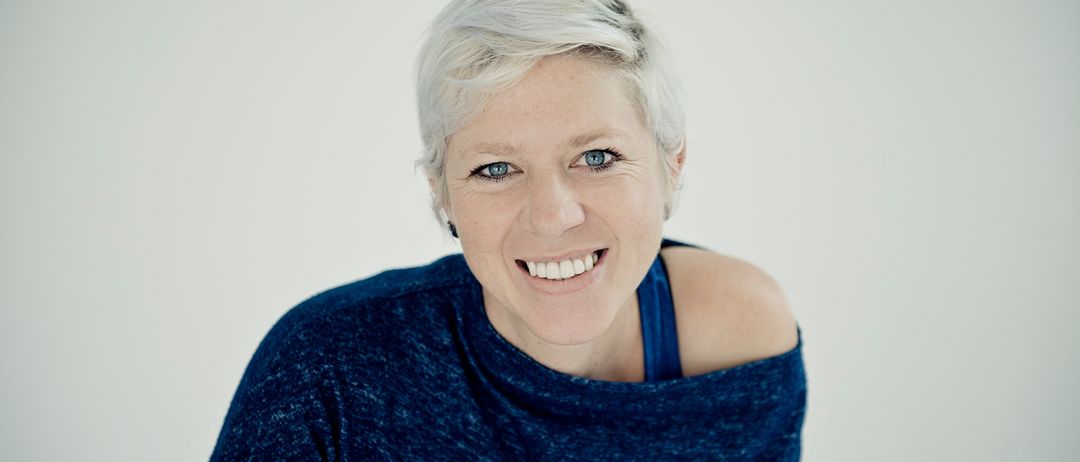 Eine Frau mit kurzen weißen Haaren und blauen Augen lächelt. Sie trägt ein blaues Off-Shoulder-Shirt. Der Hintergrund ist weiß.