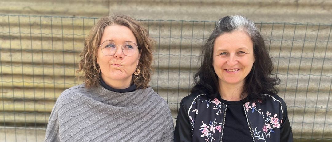 Zwei Frauen sitzen im Freien. Eine hält ein grünes Buch und die andere einen Stift. Sie sitzen vor einer Wellblechwand.