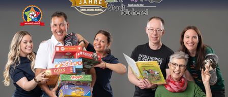 Gruppe von Menschen, die zusammenstehen, einige halten Spielzeug, Bücher und eine Puppe, mit einer Ausstellung auf einer Holzkiste vor ihnen, auf einem dunklen Hintergrund.