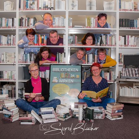 Eine Gruppe von Menschen posiert für ein Foto in einer Bibliothek mit Büchern um sie herum. Sie sitzen und stehen vor Regalen voller Bücher.