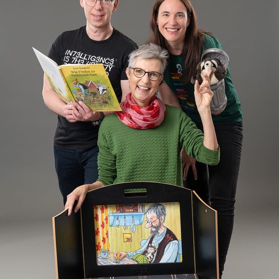 Drei Menschen stehen zusammen und posieren für ein Foto. Eine Person hält ein Buch und ein Plüschtier. Eine andere Person hält ein Buch. Sie alle lächeln.