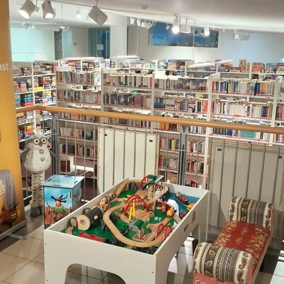 Ein Spielbereich für Kinder mit einem Spielzeug-Eisenbahnset, einer Bank und einer großen Bibliothek im Hintergrund. Die Bibliothek ist mit verschiedenen Büchern in Regalen gefüllt, und ein Balkon mit Geländer bietet einen Blick auf den Bereich. Eine Eulenstatue ist auf der linken Seite sichtbar.