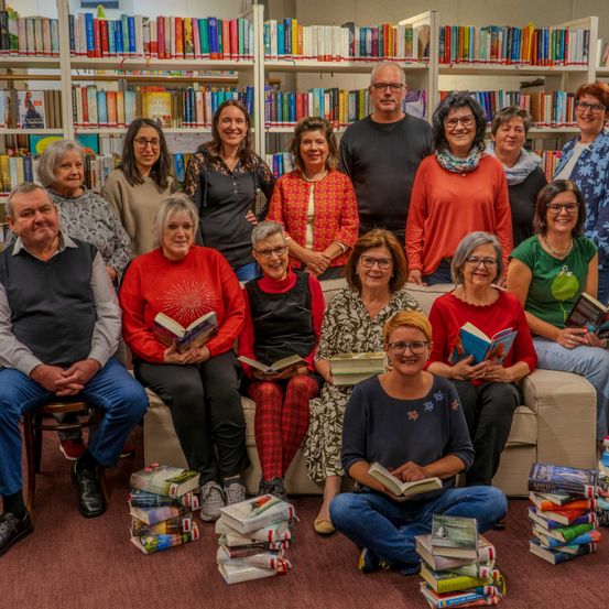 Eine Gruppe von Menschen posiert für ein Foto in einer Bibliothek, hält Bücher in den Händen und lächelt. Sie sitzen auf einer Couch und auf dem Boden. Die Bibliothek hat Regale voller Bücher.