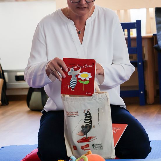 Eine Frau sitzt auf dem Boden und hält ein Buch mit dem Titel 'Was macht die Maus?' Das Buch hat eine gezeichnete Maus auf dem Cover. Sie trägt ein weißes Hemd und blaue Hosen.