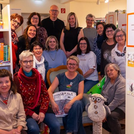 Eine Gruppe von Frauen und Männern mit Brillen posiert für ein Foto in einer Bibliothek. Die Frau in der Mitte hält ein Buch und trägt eine Schürze. Ein gedönstes Eule ist rechts.