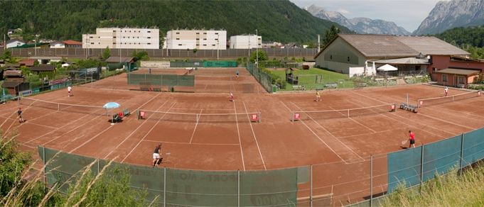 Ein Tennisplatz mit aktiven Spielern, umgeben von einem Zaun, mit Gebäuden und Bergen im Hintergrund.