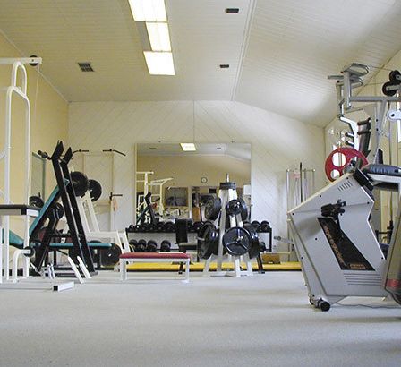Ein gut ausgestattetes Fitnessstudio mit verschiedenen Trainingsgeräten, Gewichten und einer Bank, vor einer gelben Wand mit Beleuchtung.