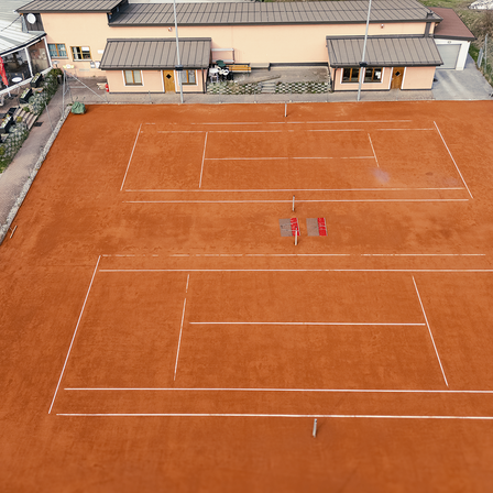Eine Draufsicht auf einen Tennisplatz mit zwei sichtbaren Plätzen, umgeben von einem Gebäude mit grauen Dächern. Die Plätze sind leer, und der Boden ist braun.