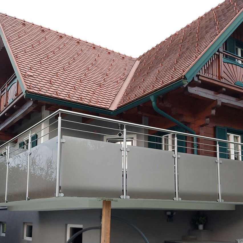 Ein Haus mit braunem Dach und grünem Rand hat einen Balkon mit Metallgeländer. Es gibt Glasscheiben am Geländer und eine Topfpflanze an der Wand darunter.