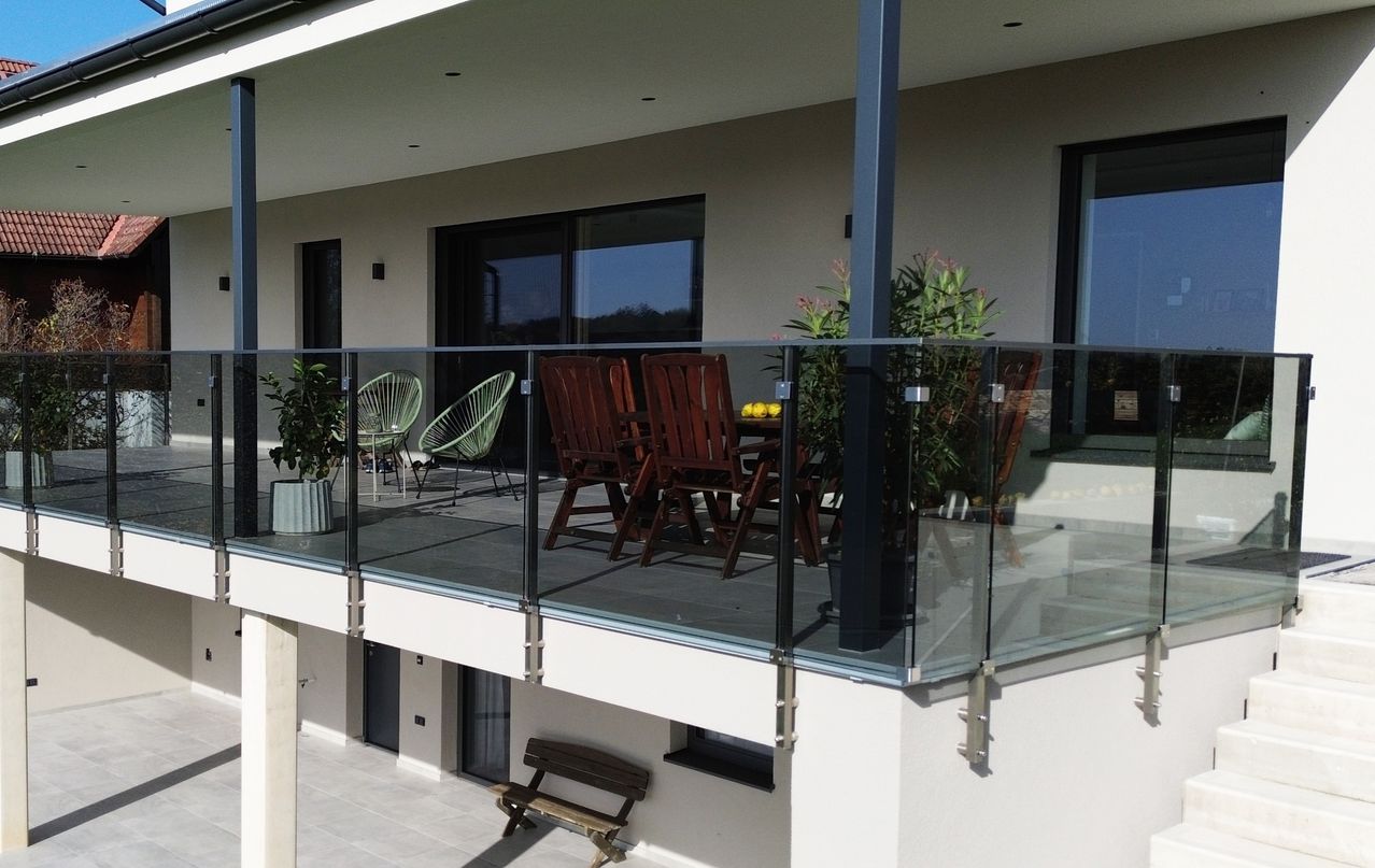 Ein Balkon mit Glasgeländer zeigt einen Essbereich mit Tisch und Stühlen. Topfpflanzen stehen auf beiden Seiten der Geländer. Der Balkon befindet sich im zweiten Stock.