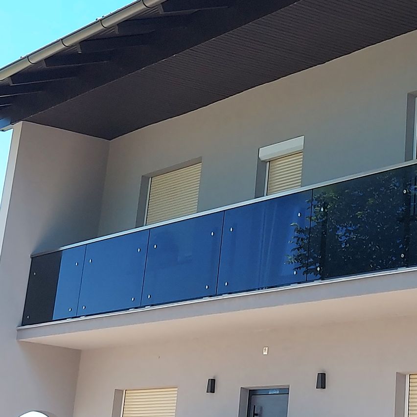 Ein Balkon mit blauen Geländern befindet sich im zweiten Stock eines Hauses. Das Haus hat weiße Wände und ein graues Dach. Zwei Fenster mit geschlossenen Jalousien sind auf dem Balkon sichtbar.