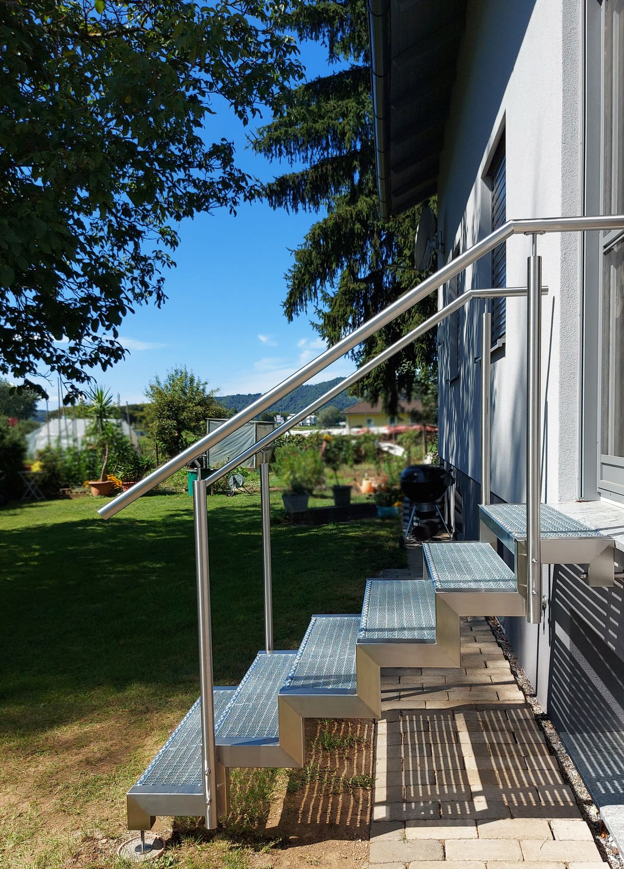 Eine Reihe moderner, rostfreier Stahlstufen mit einem Handlauf, die zu einem weißen Haus führen. Die Stufen befinden sich vor einem Rasen mit Pflanzen, einem Haus und einem Berg im Hintergrund.