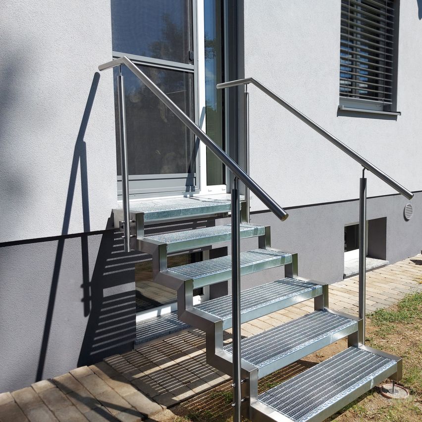 Edelstahltreppe mit drei Stufen, vor einem modernen Gebäude. Die Stufen bestehen aus einem metallischen Gitter.