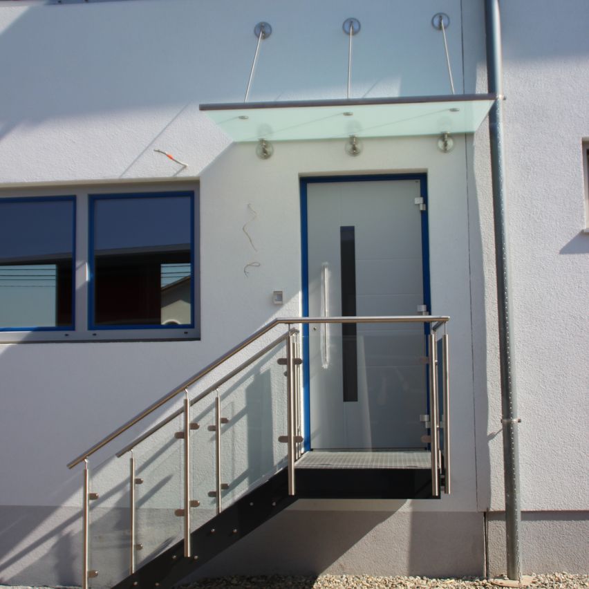 Ein modernes Hausaußen mit weißer Wand, blau gerahmten Fenstern, einer Glasüberdachung, einer weißen Tür mit blauem Rand, einem Metallgeländer und einer Treppe.