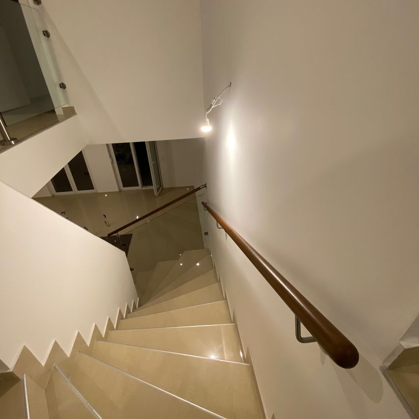 Eine moderne Treppe mit einer Glasbrüstung und einem Holzhandlauf. Die Stufen sind beige und die Wände weiß. Eine Wandlampe beleuchtet den Bereich.