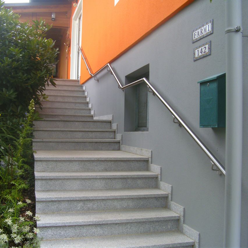 Das Bild zeigt ein graues Gebäude mit einer Treppe aus Stein, die zu einem Eingang führt. Die Stufen haben ein silbernes Geländer. Auf der linken Seite der Treppe befinden sich Pflanzen.