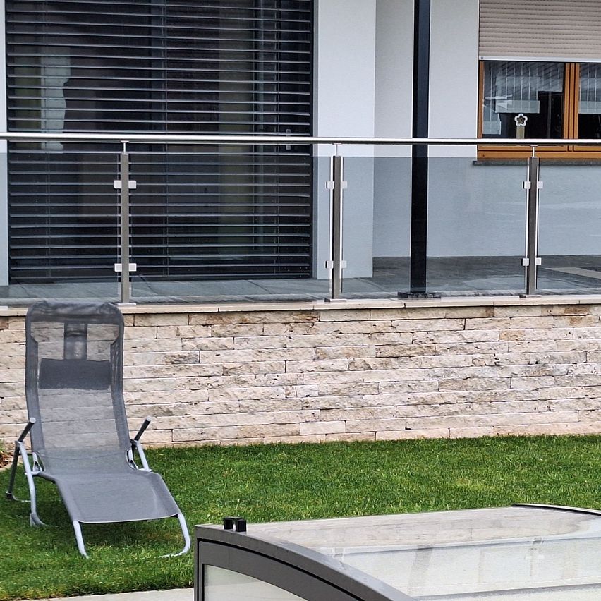 Ein Balkon mit einer Glasbrüstung und einem Liegestuhl auf einem Rasen. Der Balkon hat eine Steinmauer, ein geschlossenes Fenster und ein Fenster mit Jalousien.