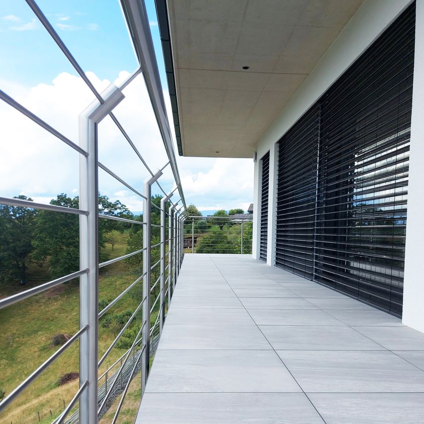Eine Außenterrasse mit grau gefliestem Boden, Metallgeländern und geschlossenen schwarzen Rolläden. Die Terrasse blickt auf ein Grasfeld mit Bäumen und einem Haus in der Ferne.