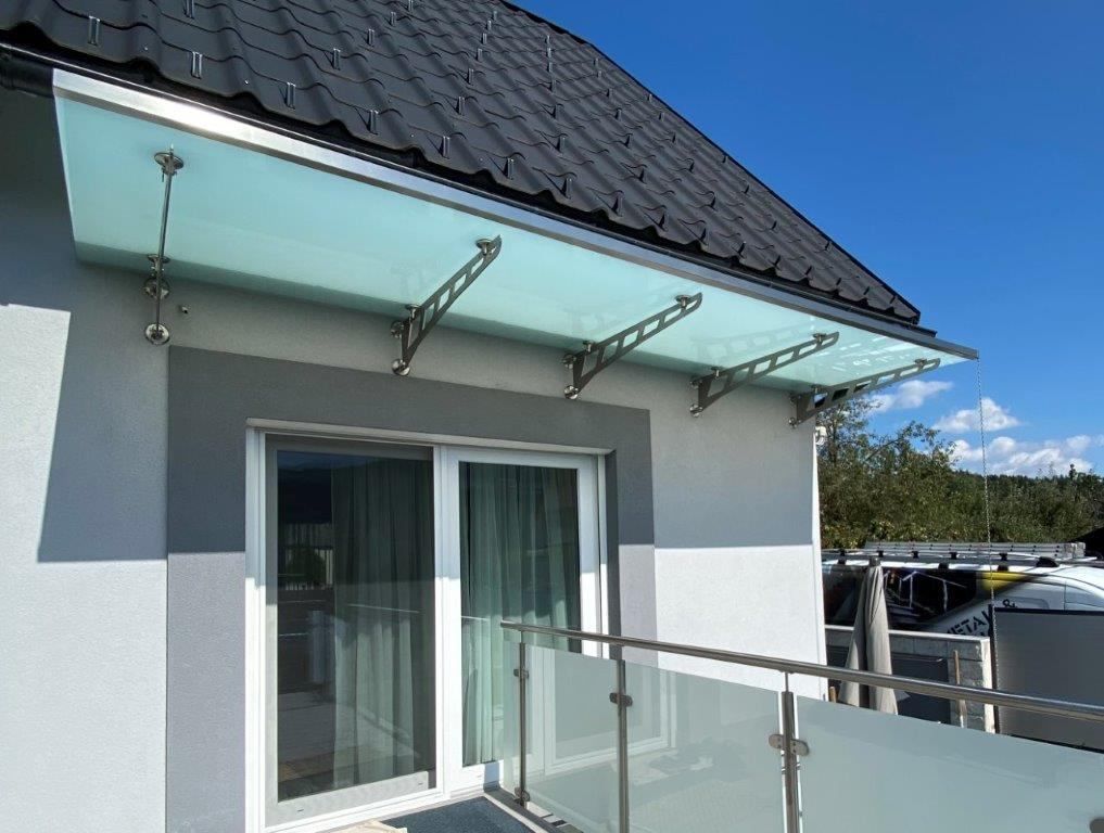 Ein Haus mit einem grauen Dach und einer Glasbalkonüberdachung. Der Balkon hat Glastüren und Geländer. Das Haus hat eine graue Wand und ein schwarzes Ziegeldach.