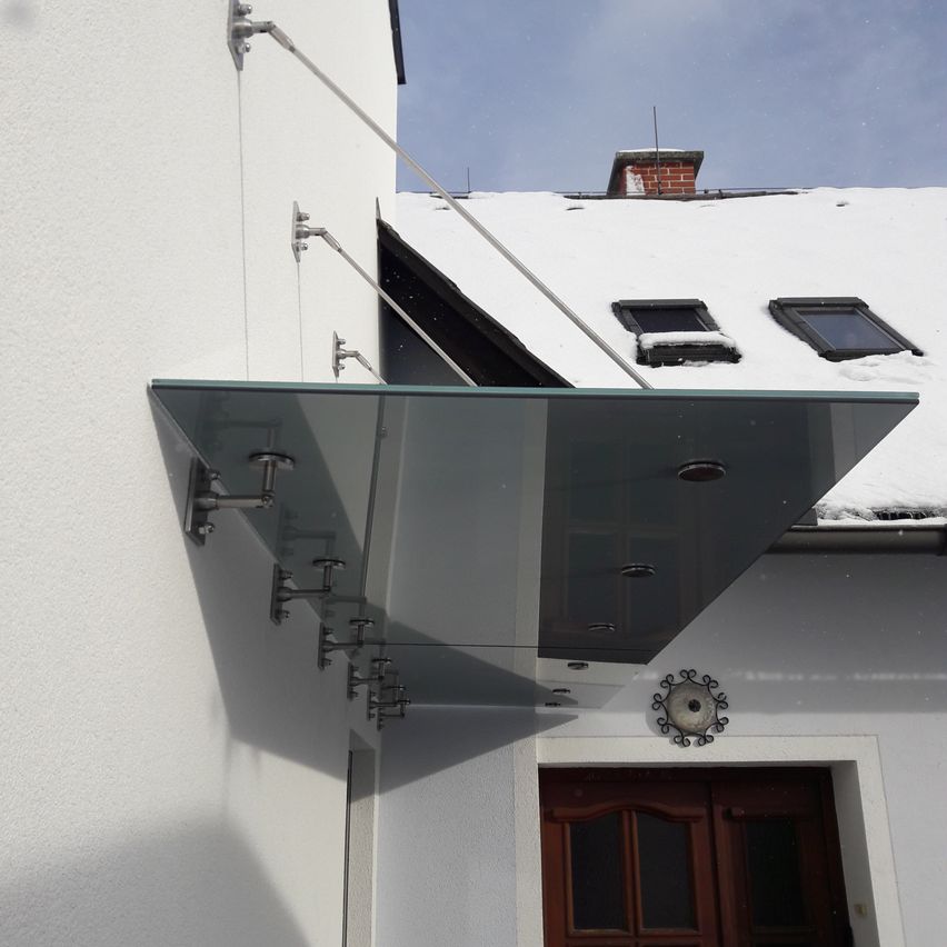 Ein modernes, glasverkleidetes Vordach mit Metallhalterungen ist an einem weißen Gebäude mit einer braunen Tür angebracht, vor einem verschneiten Hintergrund.