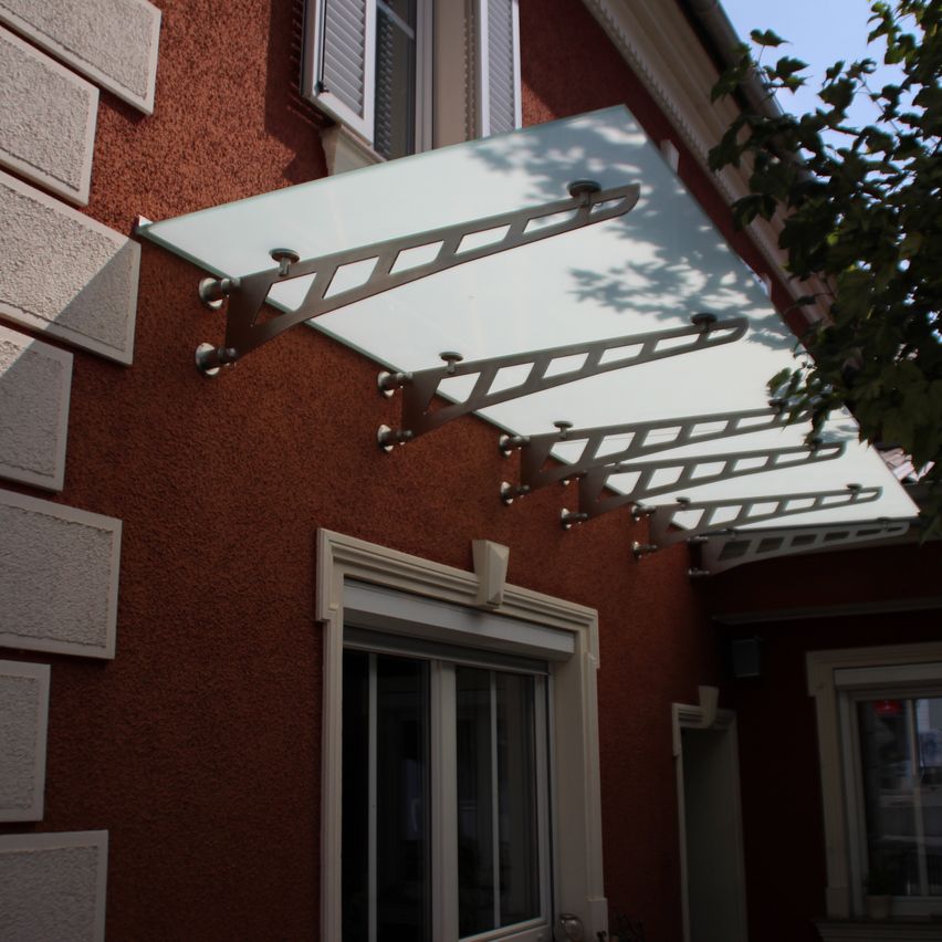 Ein rotes Gebäude hat ein weißes Vordach, das von Metallstützen gehalten wird. Darunter befindet sich eine Glastür und ein Fenster.