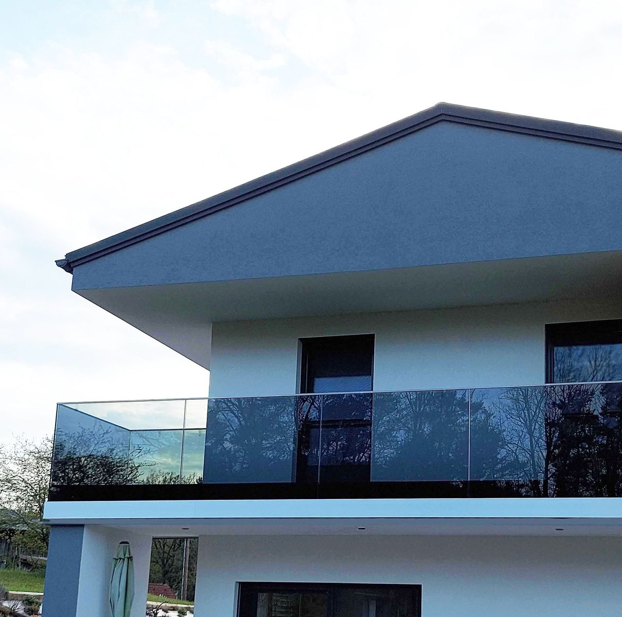 Ein modernes Haus mit einer Glasbalkon und Fenstern, umgeben von Bäumen unter einem teilweise bewölkten Himmel.