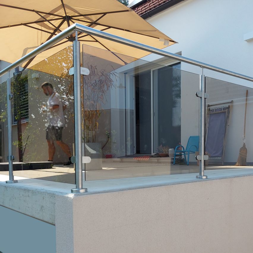 Eine Person geht auf einem Balkon mit einer Glasbrüstung und einem beigen Sonnenschirm. Ein blauer Stuhl und ein Besen sind in der Nähe.