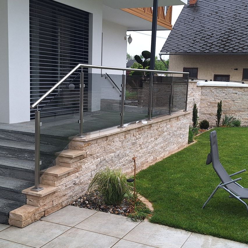 Eine Außentreppe führt zu einem modernen Haus mit einer Glasbrüstung und Steinstufen. Ein Liegestuhl steht auf dem Rasen, und eine Steinmauer umgibt das Gebiet.