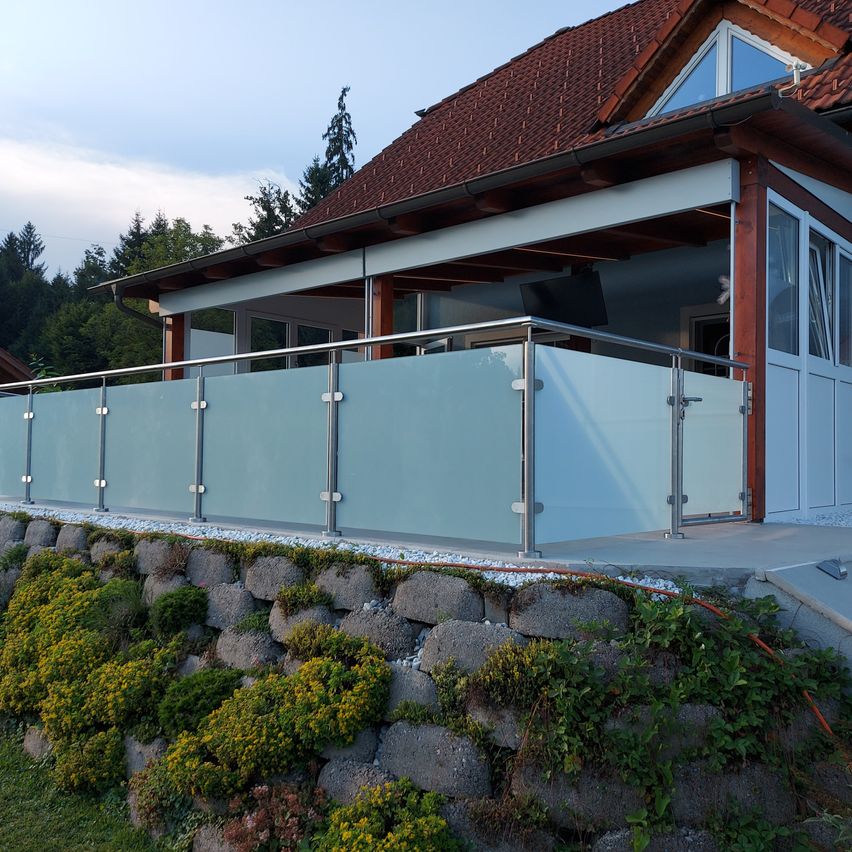 Ein modernes Haus mit einem braunen Dach, Glasfenstern und einem Balkon mit einer Glasabtrennung. Der Balkon hat ein Metallgeländer und ist von einer Steinmauer mit Pflanzen umgeben.