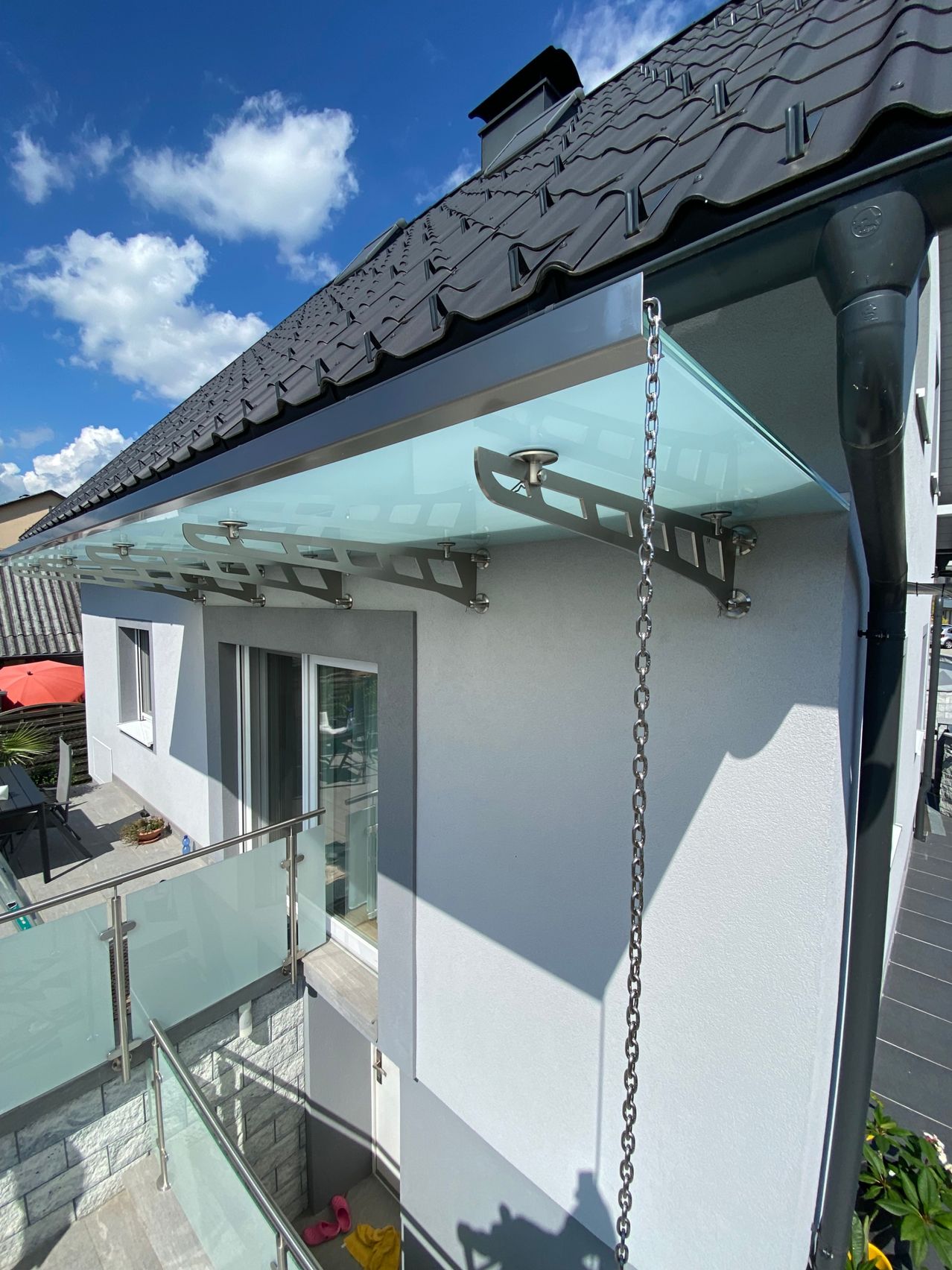 Ein Bild eines Hauses mit einer Glasüberdachung über dem Balkon, gestützt von Metallketten. Das Haus hat eine graue Außenseite und ein Ziegeldach. Der Himmel ist klar mit einigen Wolken.