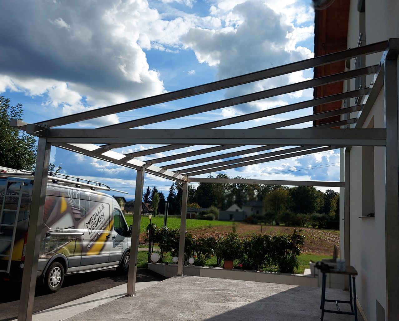 Ein Van steht unter einem Metallvordach in einem Parkplatz. Das Vordach ist an einem Gebäude mit Dach befestigt. In der Ferne befinden sich Bäume und ein Haus.