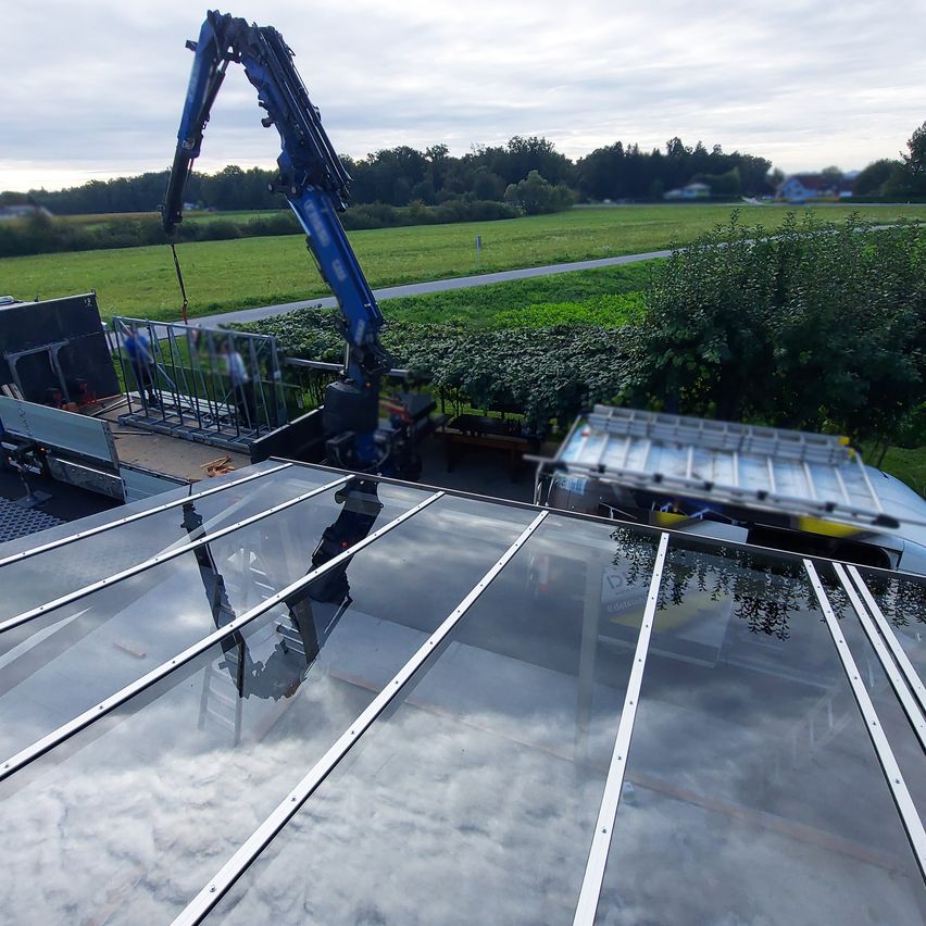 Ein blauer Kran hebt Glasplatten von einem Lkw auf ein Dach in einem ländlichen Gebiet. Arbeiter bereiten die Installation der Glasplatten vor.